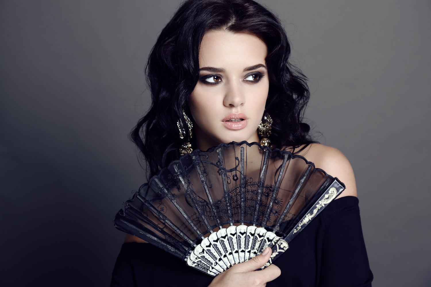 Das Bild zeigt eine elegante Frau mit dunklen, welligen Haaren, die einen aufwendig verzierten Fächer vor ihr Gesicht hält. Sie trägt auffällige Ohrringe und ein schwarzes, schulterfreies Kleid. Ihre makellose Haut und jugendliche Ausstrahlung deuten auf die Verwendung der Antifalten-Kosmetik Cellstar hin. Mit der Cellstar-Pflege kann sie das Schönheitsgeheimnis der Stars nutzen, um Falten zu reduzieren und ihre Haut jugendlich strahlend zu erhalten.