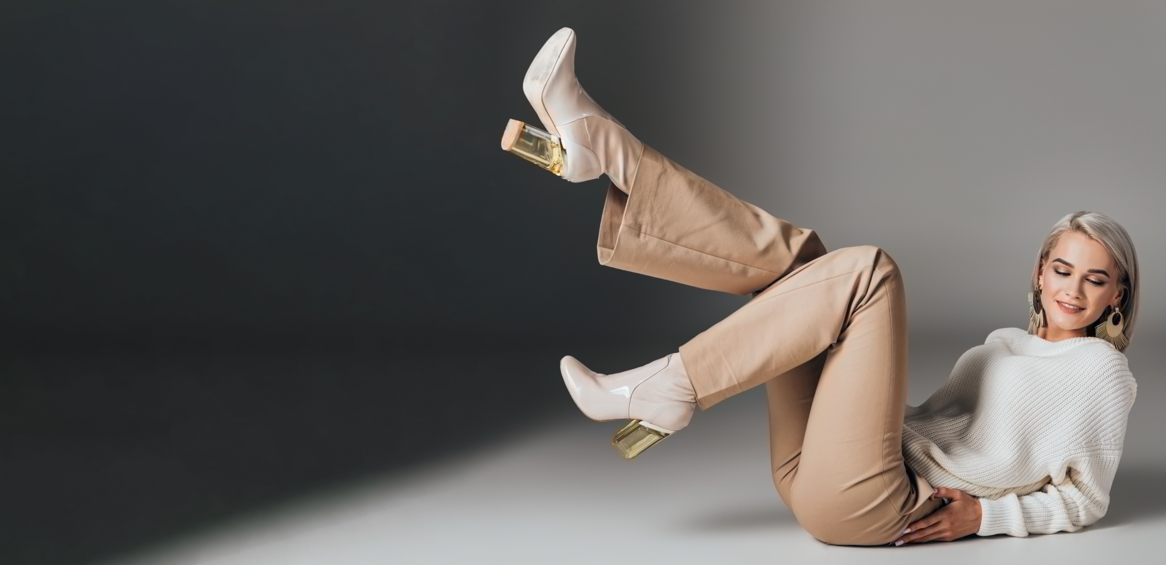 Das Bild zeigt eine Frau, die entspannt auf dem Boden liegt und dabei beige Hosen, einen weißen Pullover sowie Stiefeletten trägt. Sie fühlt sich sichtlich in ihrem jugendlichen Körper wohl, was auf die Wirkung der innovativen Shapewear "Marie - beste Freundin" zurückzuführen ist. Sie lächelt entspannt in die Kamera und ist voller Selbstbewusstsein.