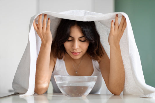 Eine junge Frau führt ein Gesichtsdampfbad durch, indem sie ihr Gesicht über eine Schale mit heißem Wasser hält und ihren Kopf mit einem weißen Handtuch abdeckt, um den Dampf zu intensivieren.